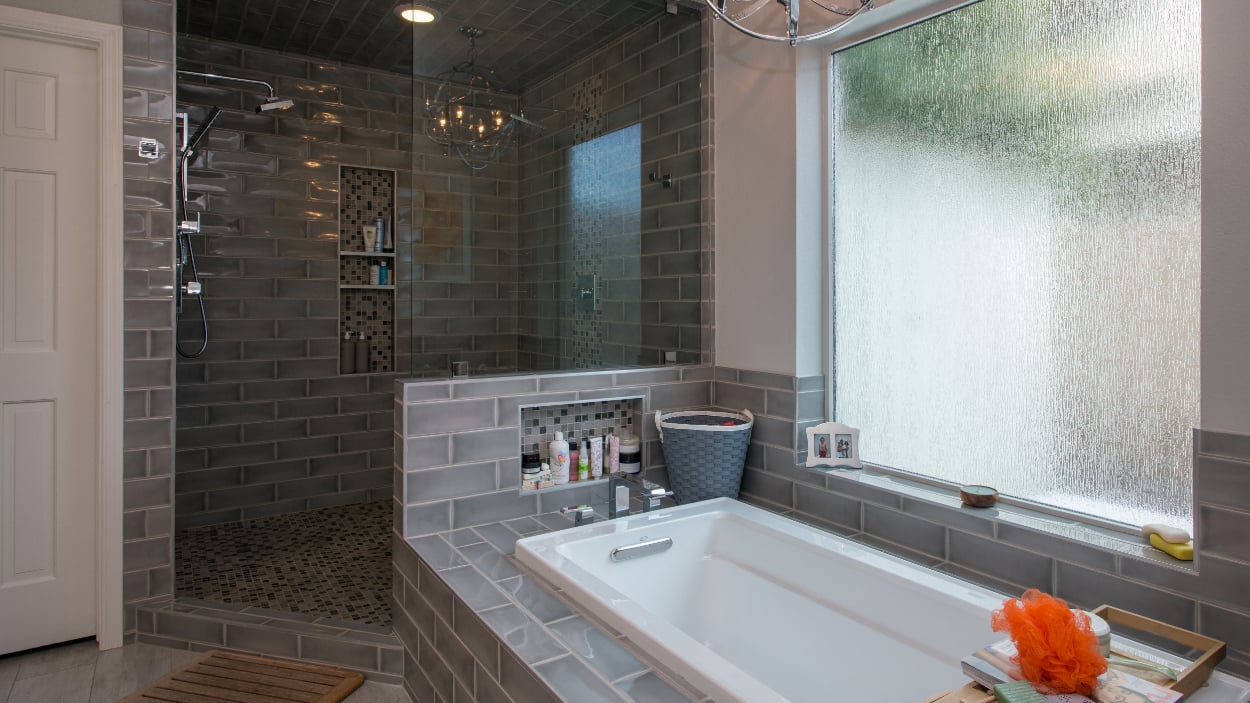 Tempe master bathroom remodel after photo showing the new doorless walk-in shower with two showerheads on the left, drop-in bathtub with rain-glass privacy window on the right, polished chrome metal globe chandelier overhead, gray Grace Grigio brickset tile throughout, and wood-look tile flooring.